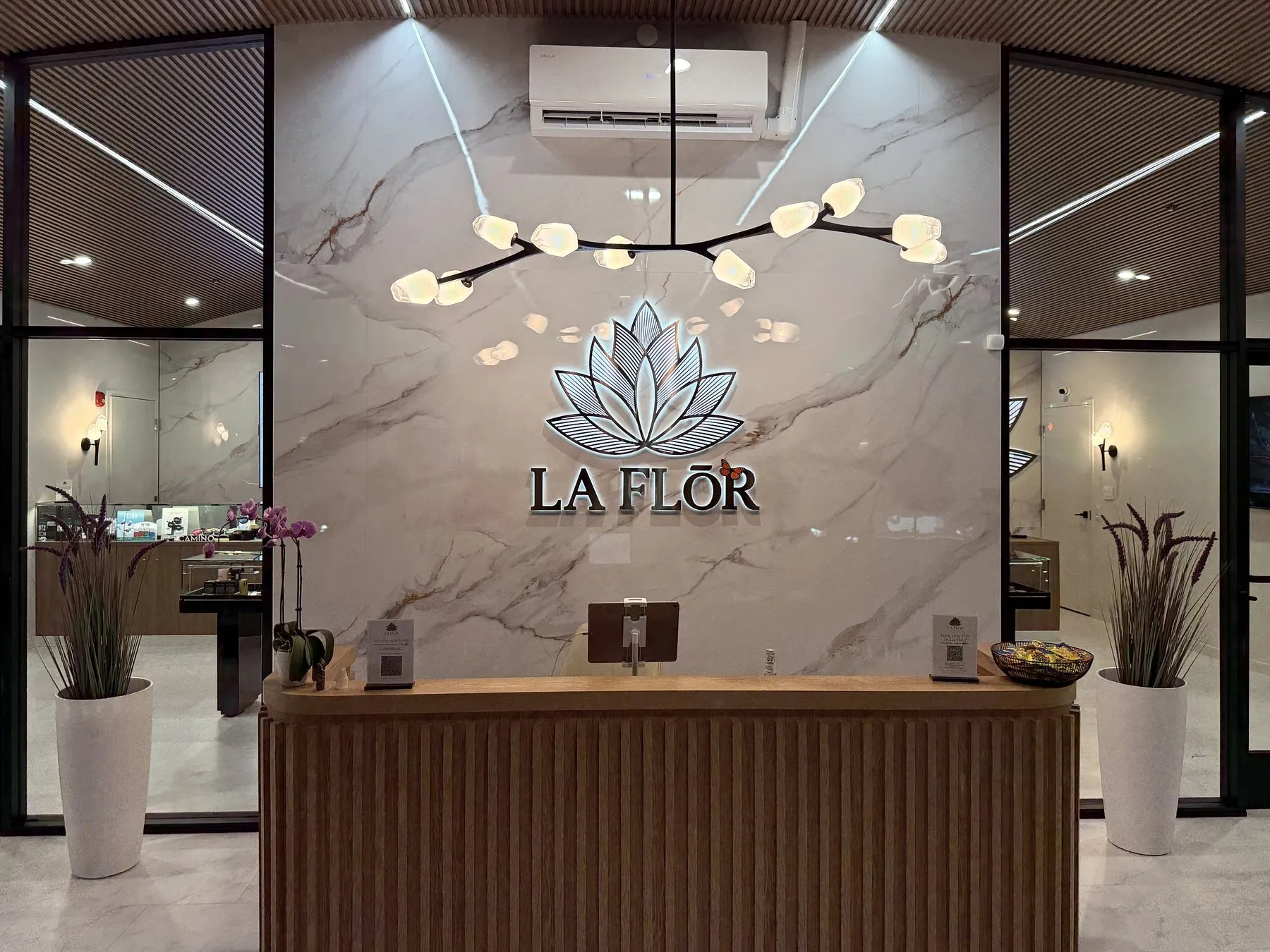 The reception desk in slatted oak, the lit lotus and LA FLŌR sign on the marble wall behind it under a branch chandelier.